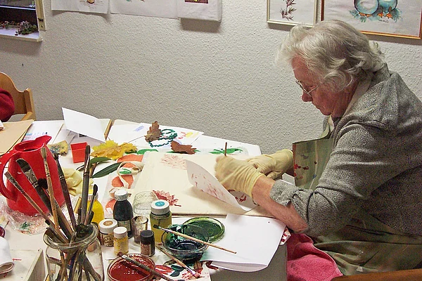 Bewohnerin beim Malen an einem Tisch voller Pinsel, Farbtöpfe und Papier