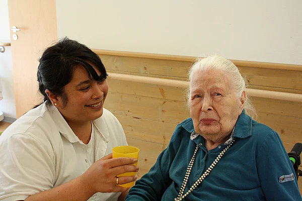 Bewohnerin neben einer Mitarbeiterin, die ihr zu Trinken gibt